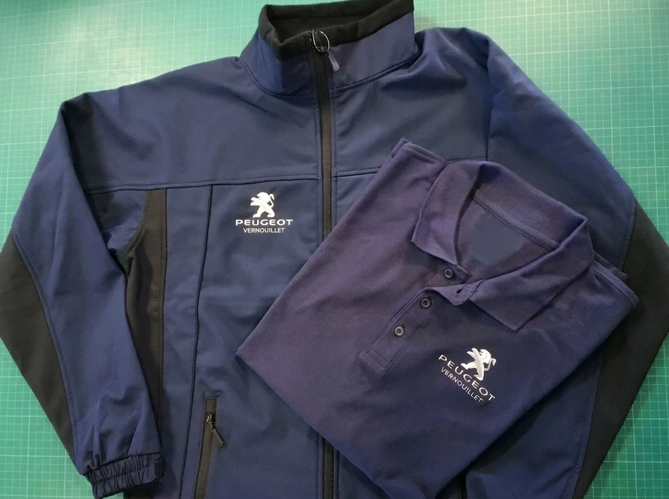 Veste softshell et polo de travail marine brodés du logo Peugeot Vernouillet, posés à plat sur un tapis de découpe vert