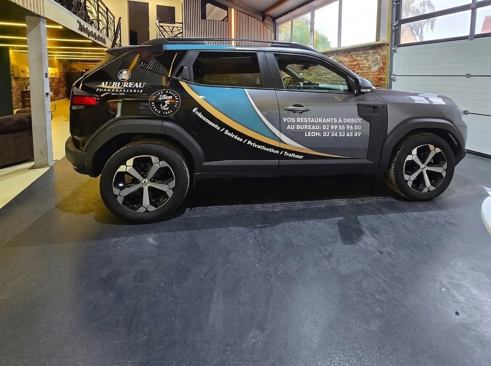 Vue rapprochée d'un utilitaire artisanal (type fourgon ou petit camion) circulant en milieu urbain ou sur route, dont la carrosserie est couverte d'un marquage publicitaire professionnel et bien visible (logos, texte, design graphique). L'image doit capturer le véhicule en action, montrant clairement comment le covering transforme le utilitaire en support de communication mobile. Prise de vue de jour, éclairage naturel, qualité professionnelle.