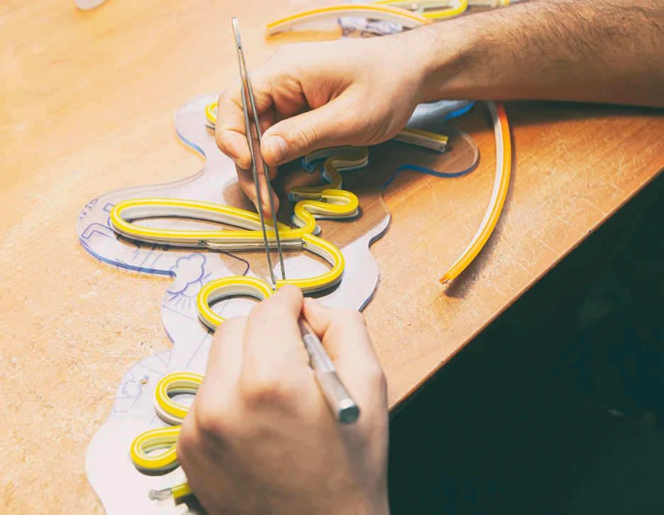 Technicien en tenue professionnelle effectuant une maintenance ou réparation d'une enseigne lumineuse LED montée en façade d'un bâtiment commercial. Le technicien est en hauteur sur un escabeau ou une nacelle, travaillant sur l'équipement d'éclairage de l'enseigne. On voit clairement le boîtier de l'enseigne LED, les câbles électriques et l'expertise technique en cours. L'image doit transmettre professionnalisme, maîtrise technique et intervention réelle sur une enseigne. Arrière-plan urbain/commercial discret pour montrer le contexte réel de l'installation.
