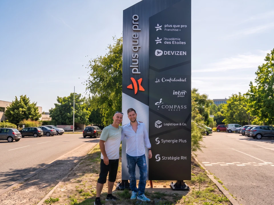 Une rue commerçante avec des magasins présentant des enseignes murales lumineuses, des totems et des bâches publicitaires visibles, capturant l'impact visuel et l'identification commerciale depuis la perspective d'un piéton ou automobiliste passant devant les vitrines. La scène doit montrer clairement comment ces supports extérieurs attirent l'attention à distance et marquent l'identité des commerces dans un environnement urbain réaliste.